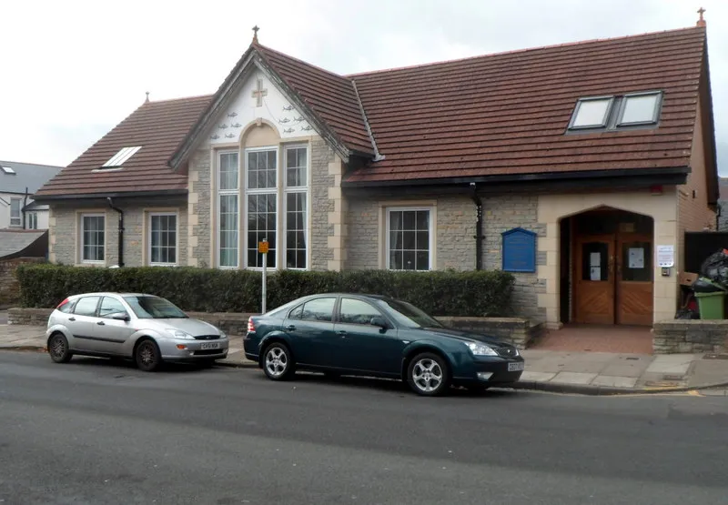 A single-storey stone and brick parish hall with a red tiled roof and arched windows, cars parked outside